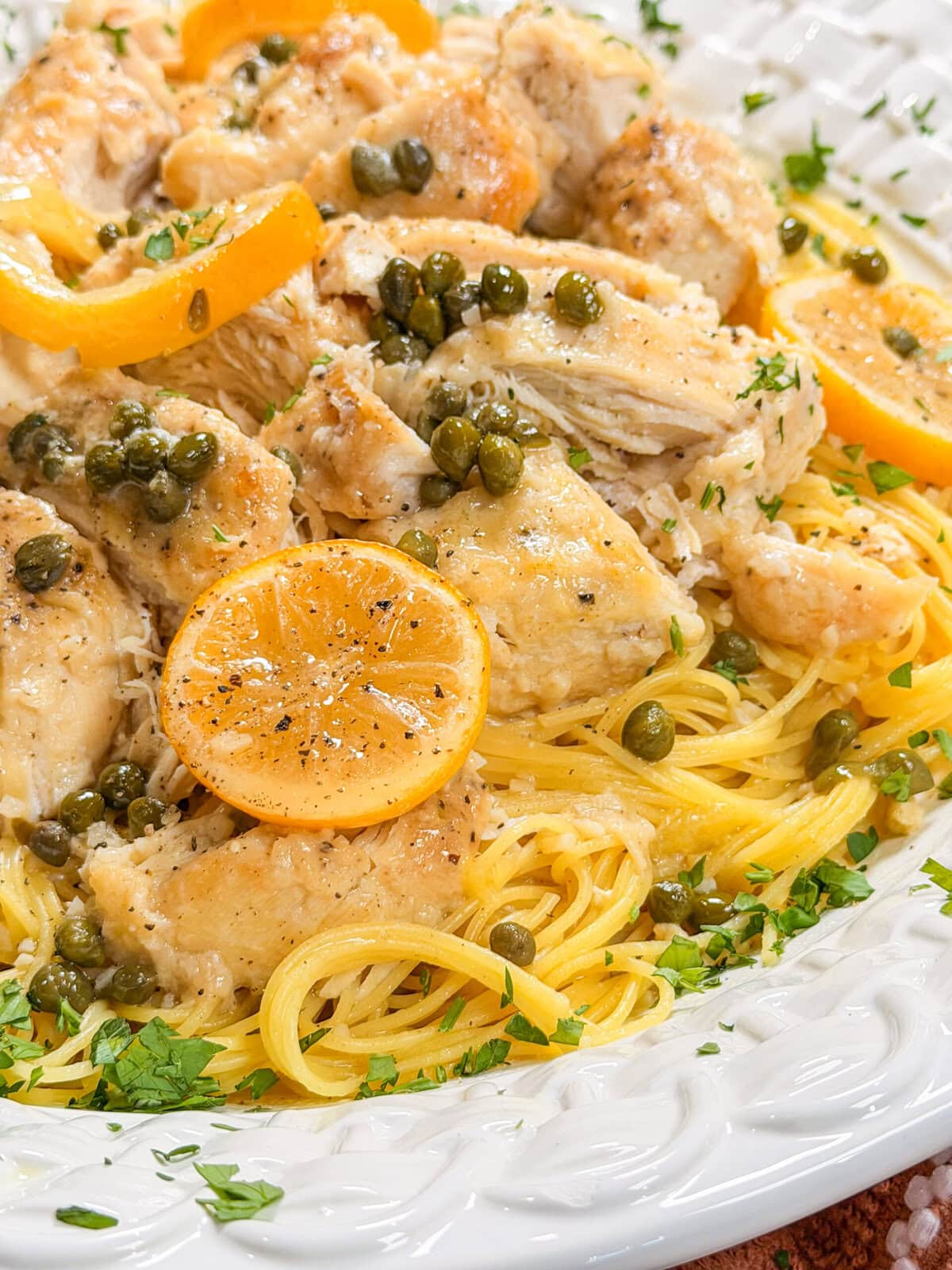 Close-up of chicken piccata served over pasta, garnished with capers, parsley, and lemon slices on a white decorative plate.