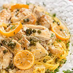 A plate of pasta topped with pieces of chicken, lemon slices, capers, and chopped parsley, served on a white decorative dish.