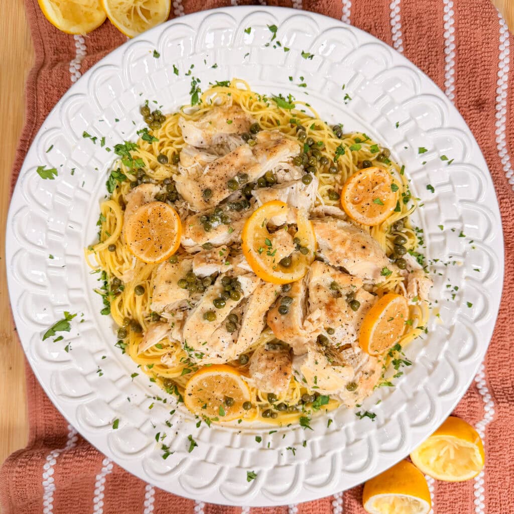 A plate of pasta topped with chicken, lemon slices, capers, and chopped herbs, on a white dish with a red towel underneath. Squeezed lemon halves are on the table.