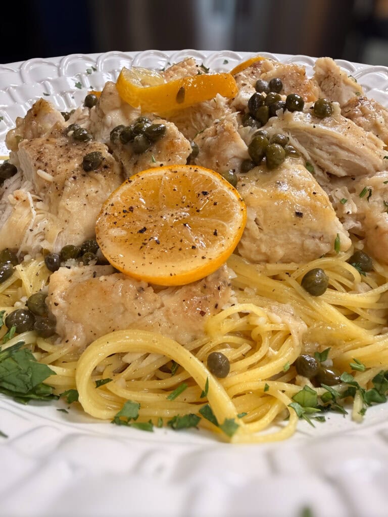 Chicken piccata with lemon slices, capers, and parsley served over spaghetti on a white plate.