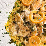 Plate of pasta topped with sliced chicken, lemon slices, capers, and chopped parsley, on a white decorative dish.