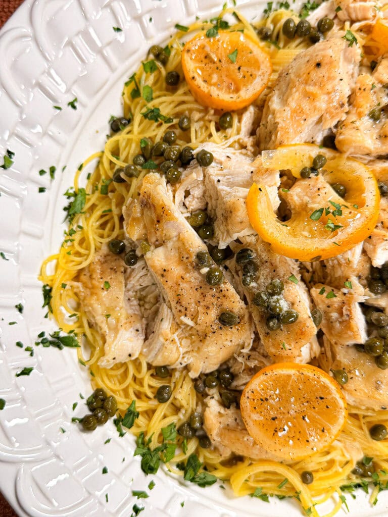 Plate of pasta topped with sliced chicken, lemon slices, capers, and chopped parsley, on a white decorative dish.