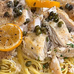 Close-up of chicken piccata served over spaghetti, garnished with capers, lemon slices, fresh herbs, and cracked black pepper.