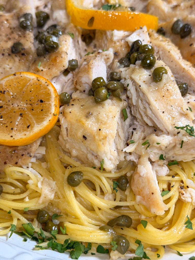 Close-up of chicken piccata served over spaghetti, garnished with capers, lemon slices, fresh herbs, and cracked black pepper.