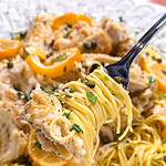 A fork lifts a bite of pasta with chicken, capers, lemon slices, and herbs from a white plate of chicken piccata.