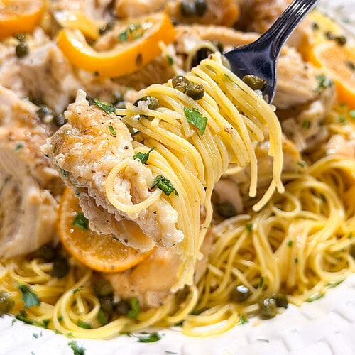 A fork lifts a bite of pasta with chicken, capers, lemon slices, and herbs from a white plate of chicken piccata.