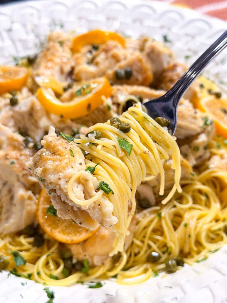 A fork lifts a bite of pasta with chicken, capers, lemon slices, and herbs from a white plate of chicken piccata.