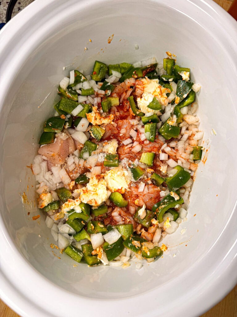 Chopped onions, green bell peppers, spices, and uncooked chicken breast in a white slow cooker, ready to be cooked.