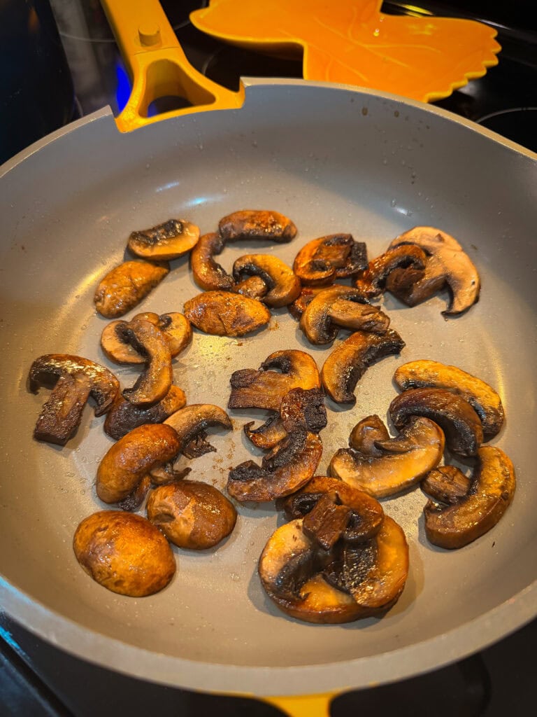 Sliced mushrooms being sautéed in a gray nonstick pan with a yellow handle on a stove, some pieces appear browned and cooked.