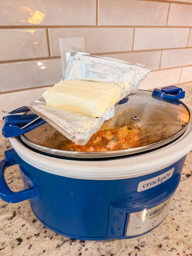 A block of cream cheese sits on top of a blue crockpot covered by its lid, with a meal cooking inside. The crockpot is on a speckled countertop in a kitchen.