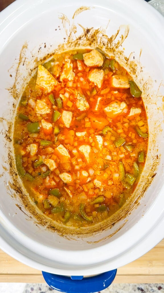 A close-up of a white slow cooker filled with a stew containing chunks of meat, diced celery, onions, and an orange-colored broth. The sides of the cooker show some splashes and seasoning residue.