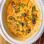 Creamy pasta with bowtie noodles, mushrooms, and green herbs in a white slow cooker, sitting on a wooden surface next to a brown striped towel.