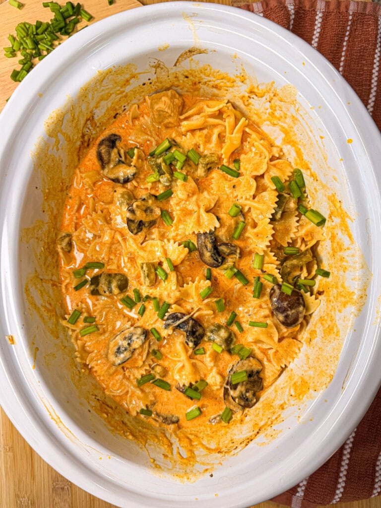 Creamy pasta with bowtie noodles, mushrooms, and green herbs in a white slow cooker, sitting on a wooden surface next to a brown striped towel.
