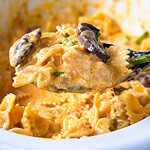 A close-up of a spoonful of creamy bowtie pasta with mushrooms, held above a white dish filled with more pasta.