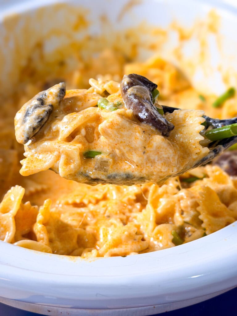 A close-up of a spoonful of creamy bowtie pasta with mushrooms, held above a white dish filled with more pasta.