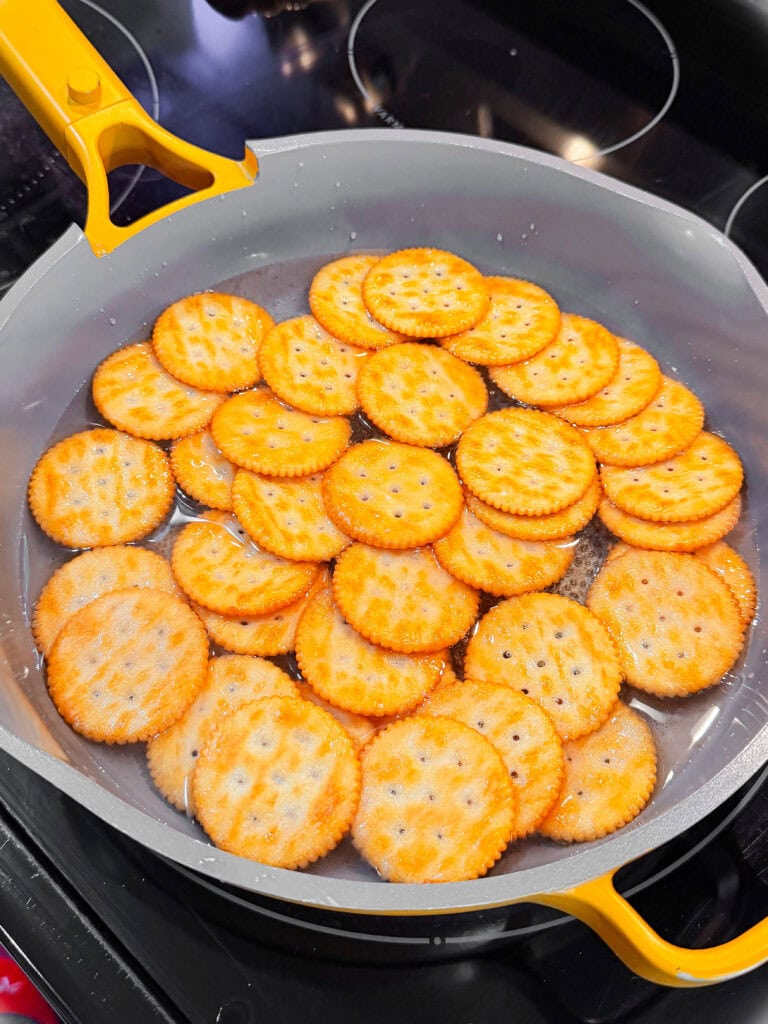 A frying pan on a stovetop filled with several round, golden-brown crackers arranged in a single layer, soaking in liquid.