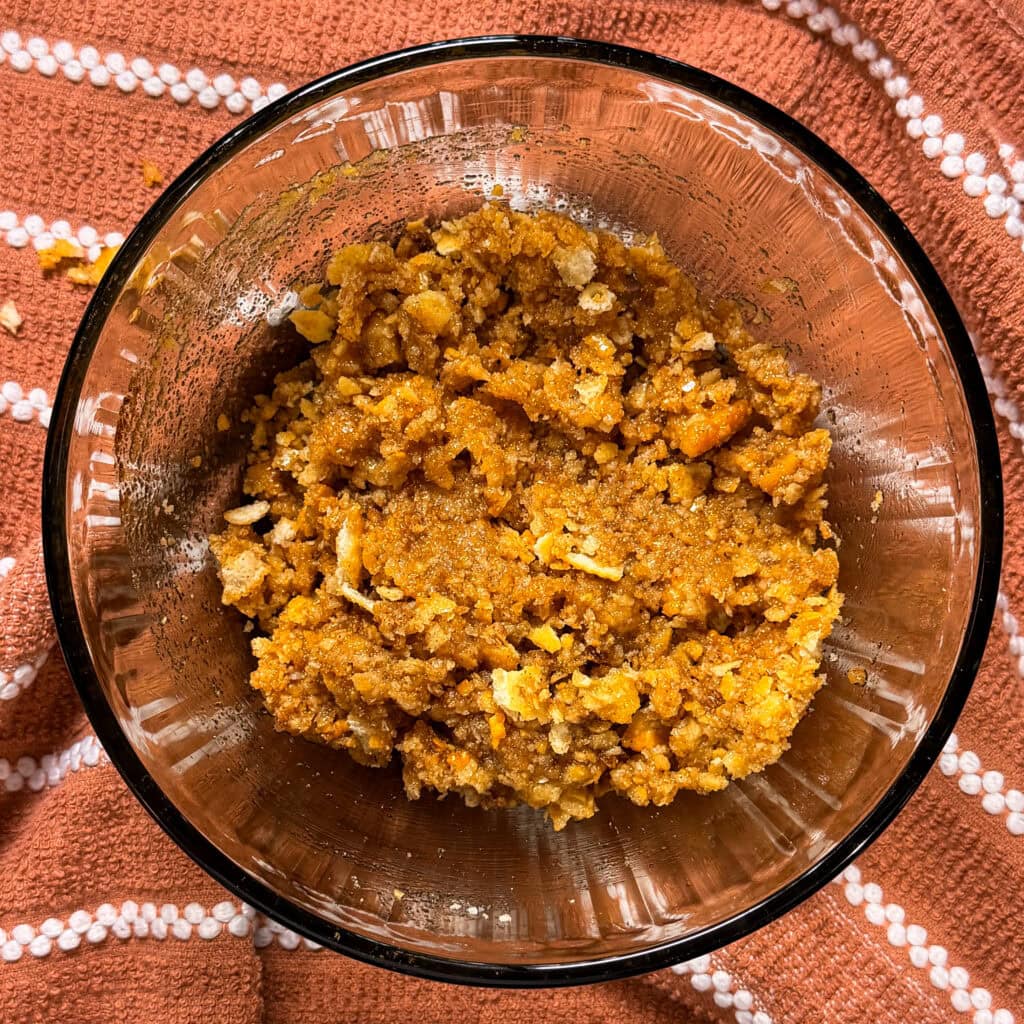 A glass bowl filled with a crumbly, orange-brown mixture sits on an orange textured cloth with white stripes. The mixture appears to contain crumbs and small flakes, resembling a dessert or snack preparation.