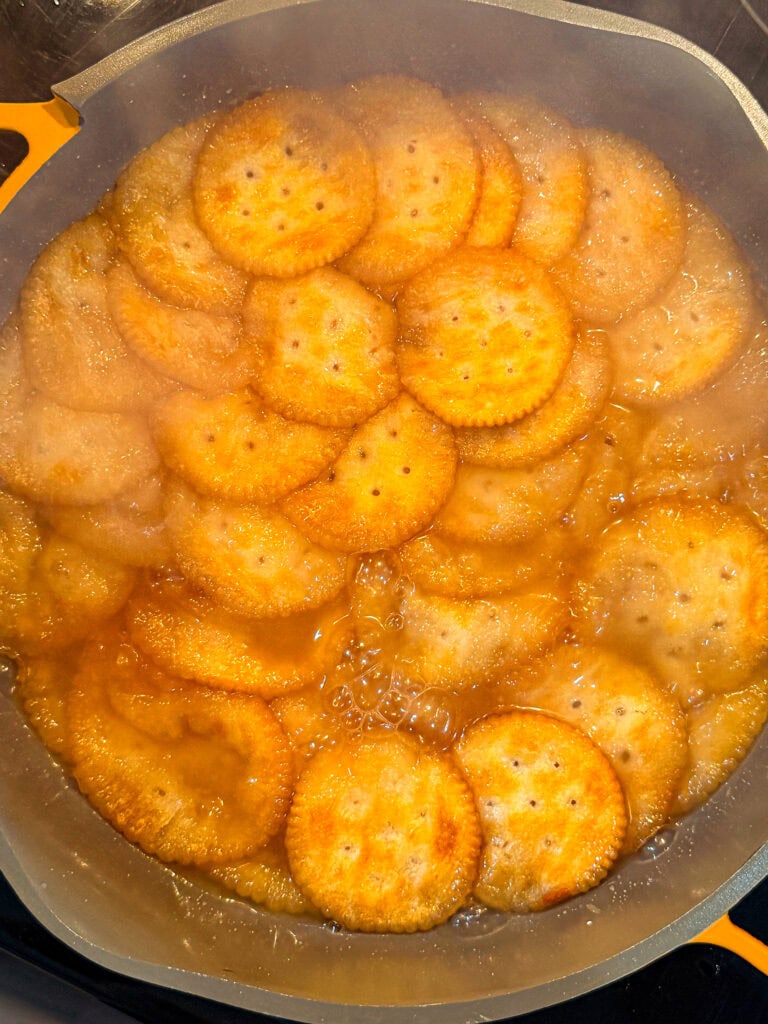 Round crackers are simmering in a pan with a bubbly, golden-brown sauce. Steam rises from the pan, and the crackers appear partially soaked and softening in the liquid.