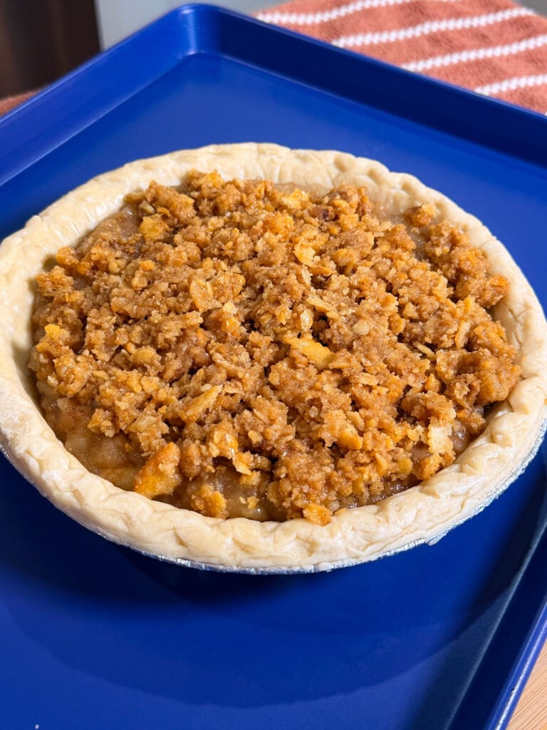 A small pie with a crumbly, golden-brown streusel topping sits on a blue tray. Theres a striped cloth visible in the background.