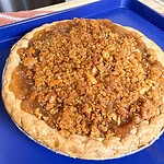 A homemade pie with a golden crumble topping sits on a blue baking sheet, cooling on a kitchen counter.