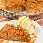 A slice of apple pie with crumb topping and a scoop of vanilla ice cream drizzled with caramel sauce on a white plate, with the rest of the pie in a glass dish behind.