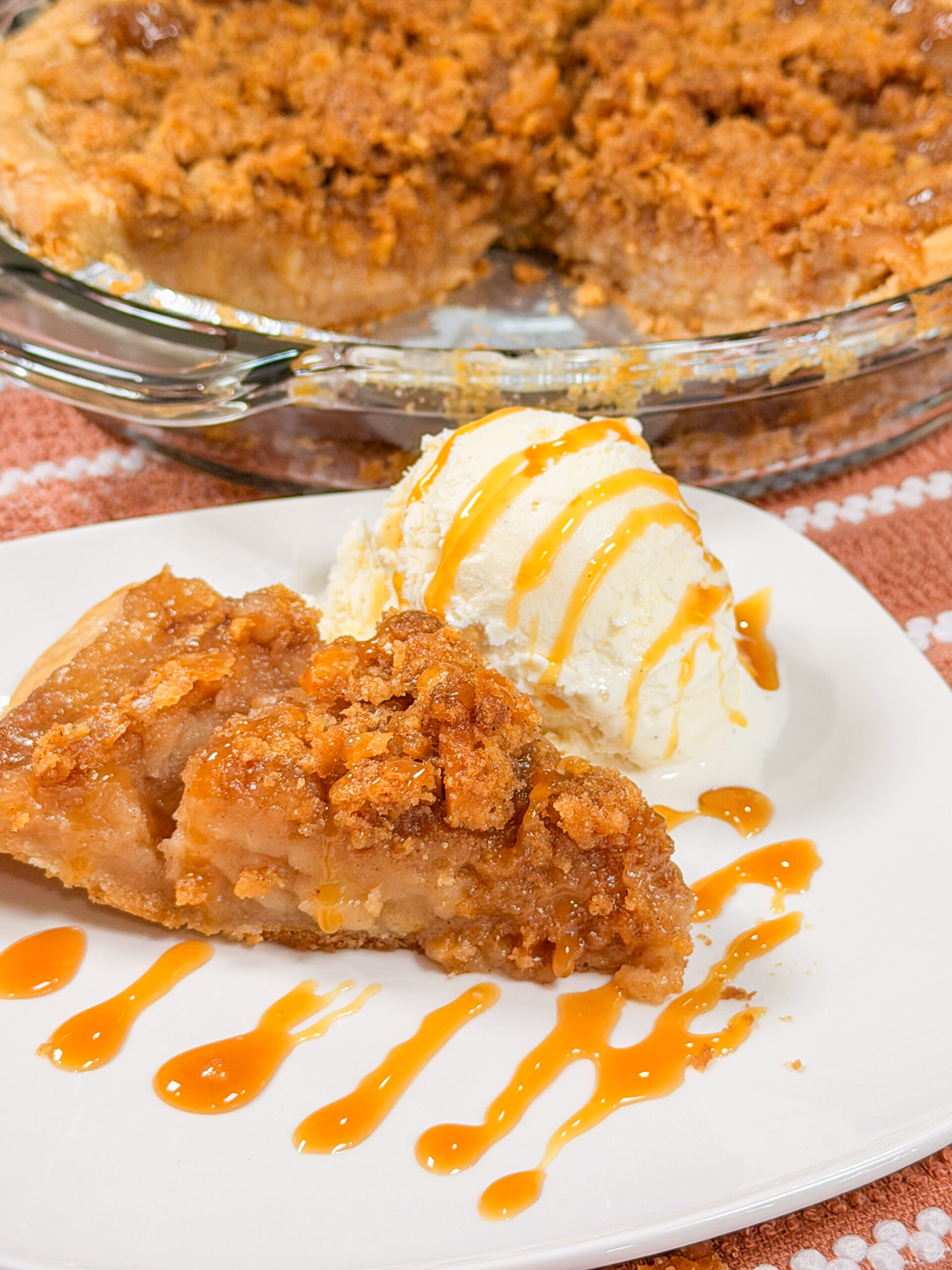 A slice of apple pie with crumb topping and a scoop of vanilla ice cream drizzled with caramel sauce on a white plate, with the rest of the pie in a glass dish behind.