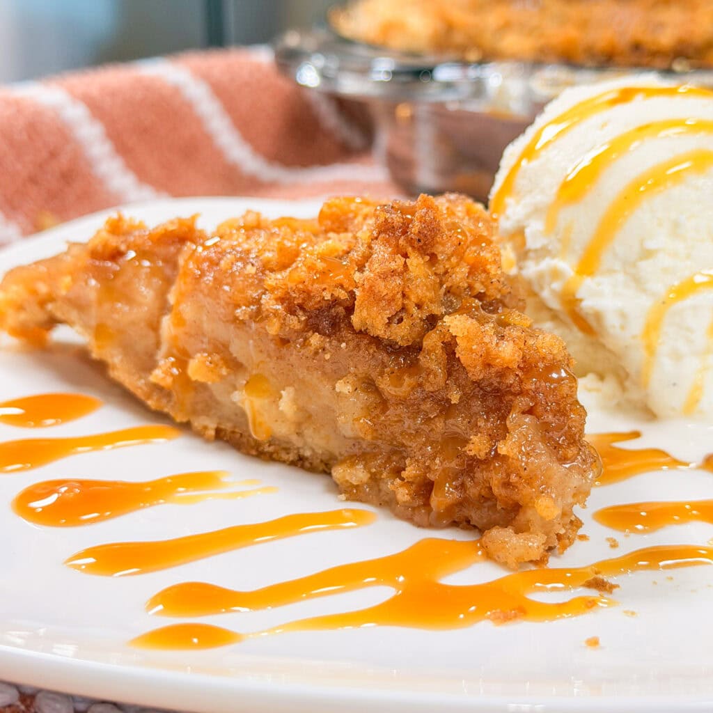 A slice of apple pie with a crumb topping is served with a scoop of vanilla ice cream, both drizzled with caramel sauce, on a white plate.