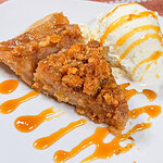 A slice of crumb-topped apple pie with a scoop of vanilla ice cream, both drizzled with caramel sauce, on a white plate.