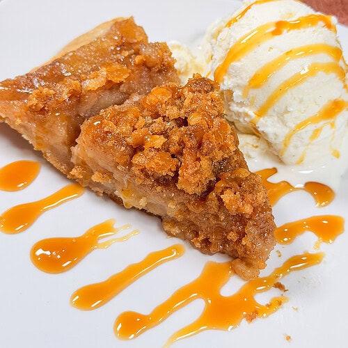 A slice of crumb-topped apple pie with a scoop of vanilla ice cream, both drizzled with caramel sauce, on a white plate.