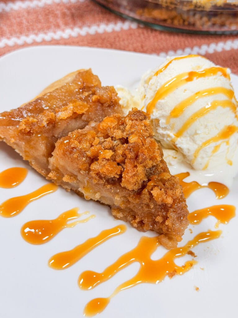 A slice of crumb-topped apple pie with a scoop of vanilla ice cream, both drizzled with caramel sauce, on a white plate.