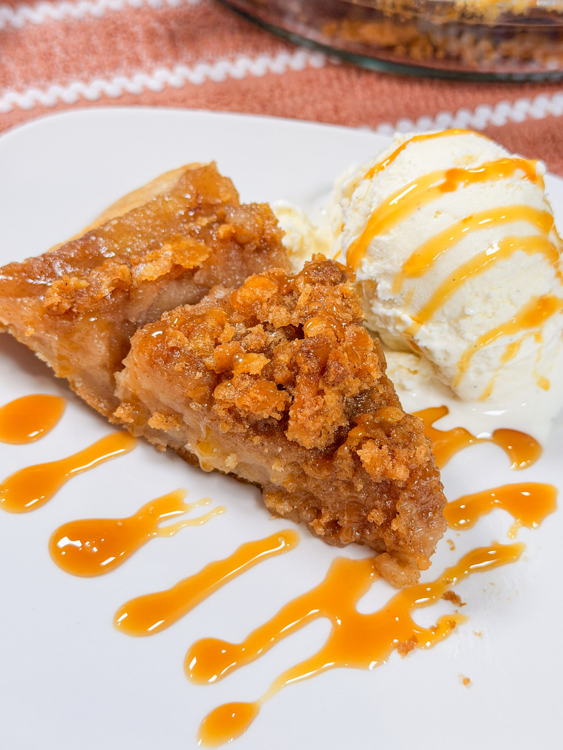 A slice of crumb-topped apple pie with a scoop of vanilla ice cream, both drizzled with caramel sauce, on a white plate.
