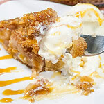 A fork holds a bite of apple crisp pie with vanilla ice cream and caramel drizzle, in front of a plate with more pie and ice cream, and a pie dish in the background.
