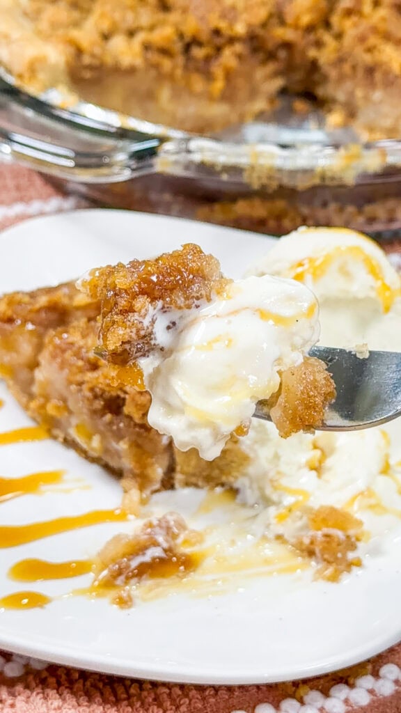 A fork holds a bite of apple crisp pie with vanilla ice cream and caramel drizzle, in front of a plate with more pie and ice cream, and a pie dish in the background.