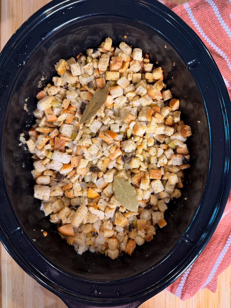 Crockpot filled with bread stuffing cubes, sprinkled with herbs and topped with two bay leaves, sits on a wooden surface next to a folded pink striped towel.