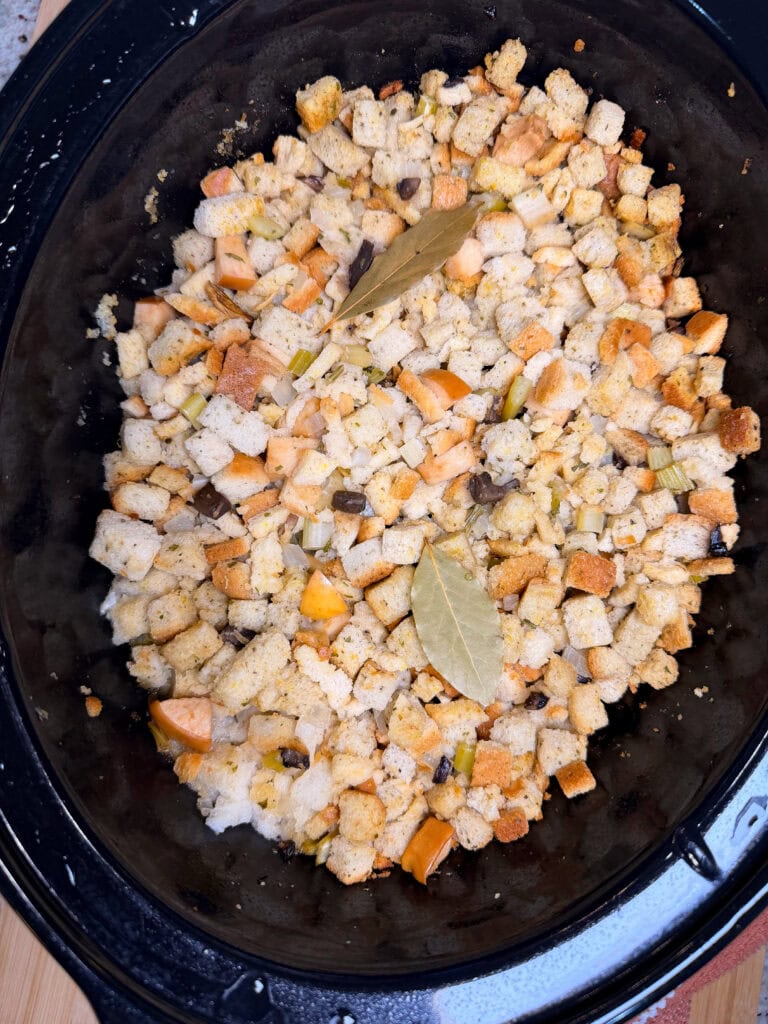 Crockpot filled with cubed bread stuffing, mixed with chopped vegetables and two bay leaves on top, ready to be cooked.