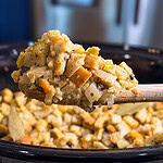 A wooden spoon holds a scoop of bread stuffing with vegetables above a slow cooker filled with more stuffing.