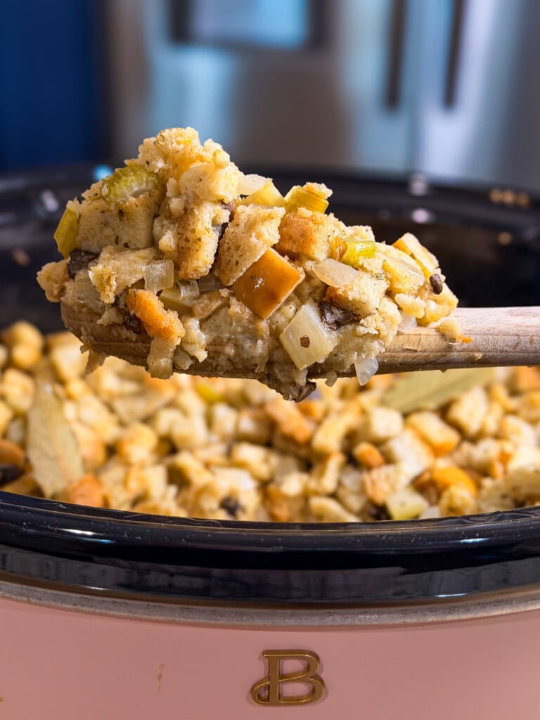 A wooden spoon holds a scoop of bread stuffing with vegetables above a slow cooker filled with more stuffing.