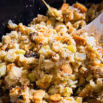 Close-up of homemade stuffing with bread cubes, celery, onions, and herbs being mixed with a wooden spoon in a bowl.