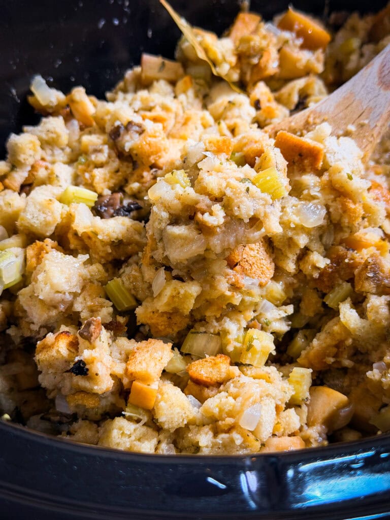 Close-up of homemade stuffing with bread cubes, celery, onions, and herbs being mixed with a wooden spoon in a bowl.