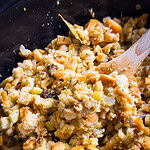Close-up of bread stuffing with chopped celery and onions in a black pot, being stirred with a wooden spoon. The stuffing appears golden and moist with visible herbs and vegetables.