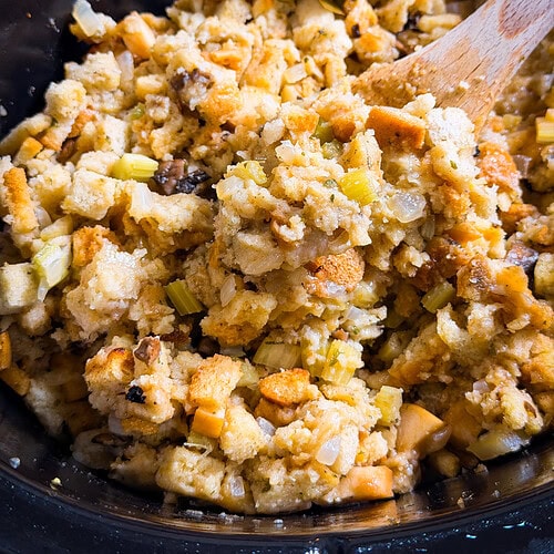 Close-up of bread stuffing with chopped celery and onions in a black pot, being stirred with a wooden spoon. The stuffing appears golden and moist with visible herbs and vegetables.