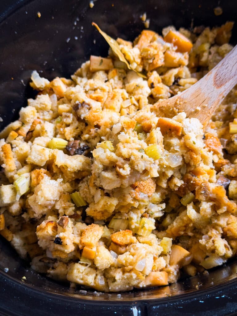 Close-up of bread stuffing with chopped celery and onions in a black pot, being stirred with a wooden spoon. The stuffing appears golden and moist with visible herbs and vegetables.