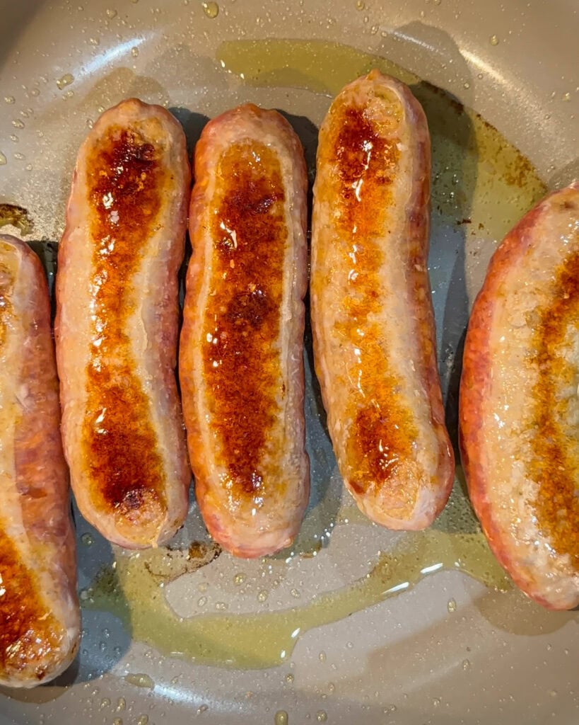 Five sausages are cooking in a pan with oil, showing browned and slightly crispy tops, indicating they are being fried or sautéed.