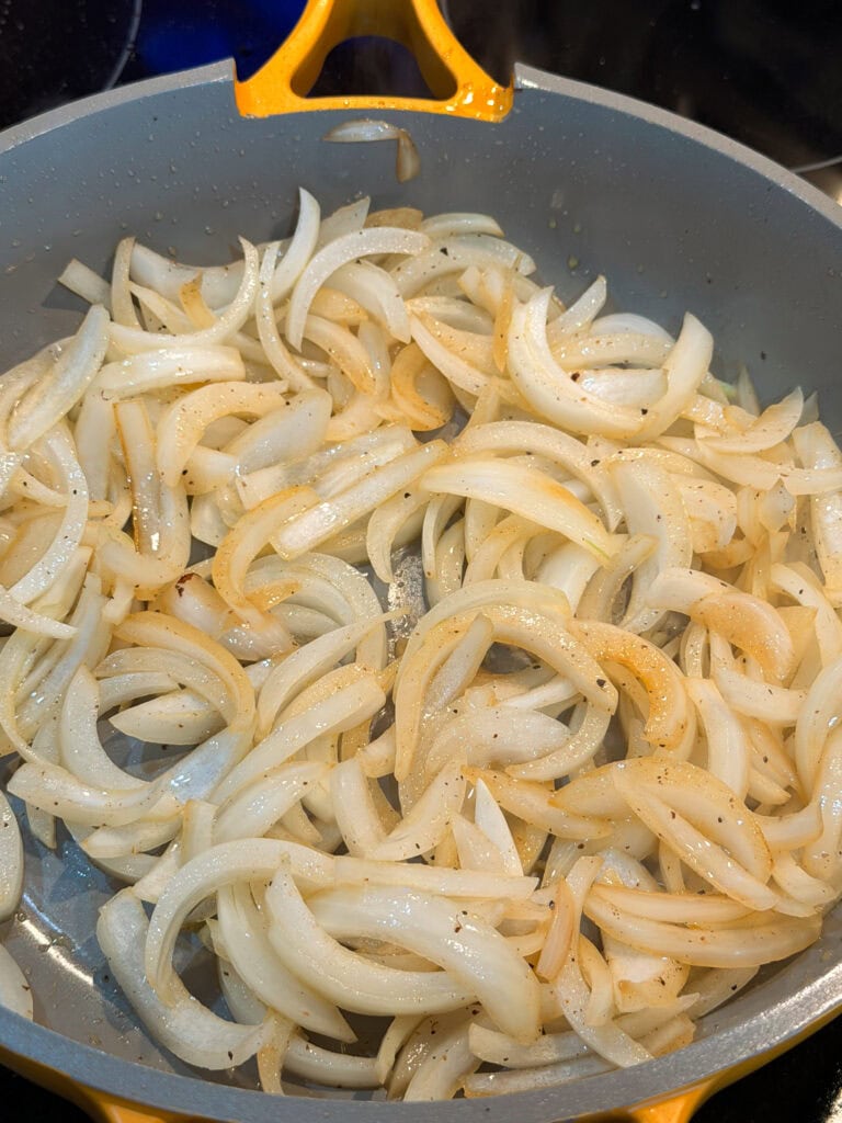 Sliced onions cooking in a gray skillet with a yellow handle. The onions are starting to soften and turn slightly golden, with visible seasoning.
