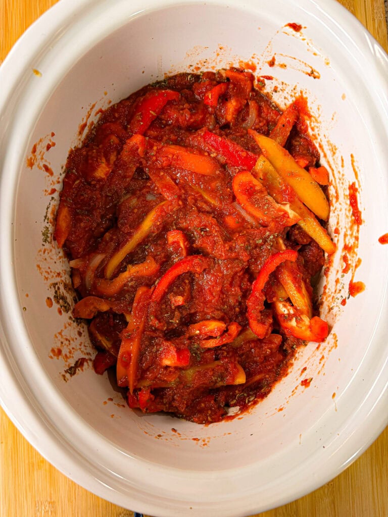 Bell pepper slices and onions in tomato sauce inside a white slow cooker, sitting on a wooden surface.