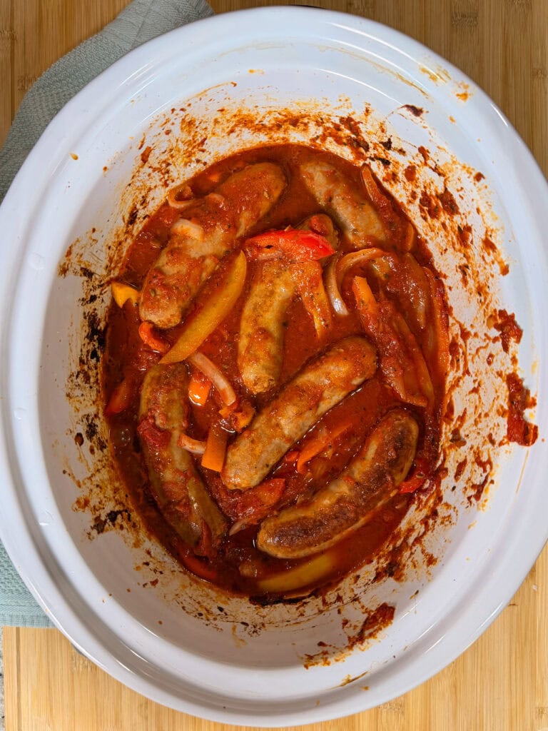 Sausages cooked in tomato sauce with sliced red and yellow peppers and onions in a white slow cooker on a wooden surface.