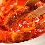 A pair of tongs holds a cooked sausage over a pot of sausages simmering in a tomato-based sauce with sliced peppers and onions.