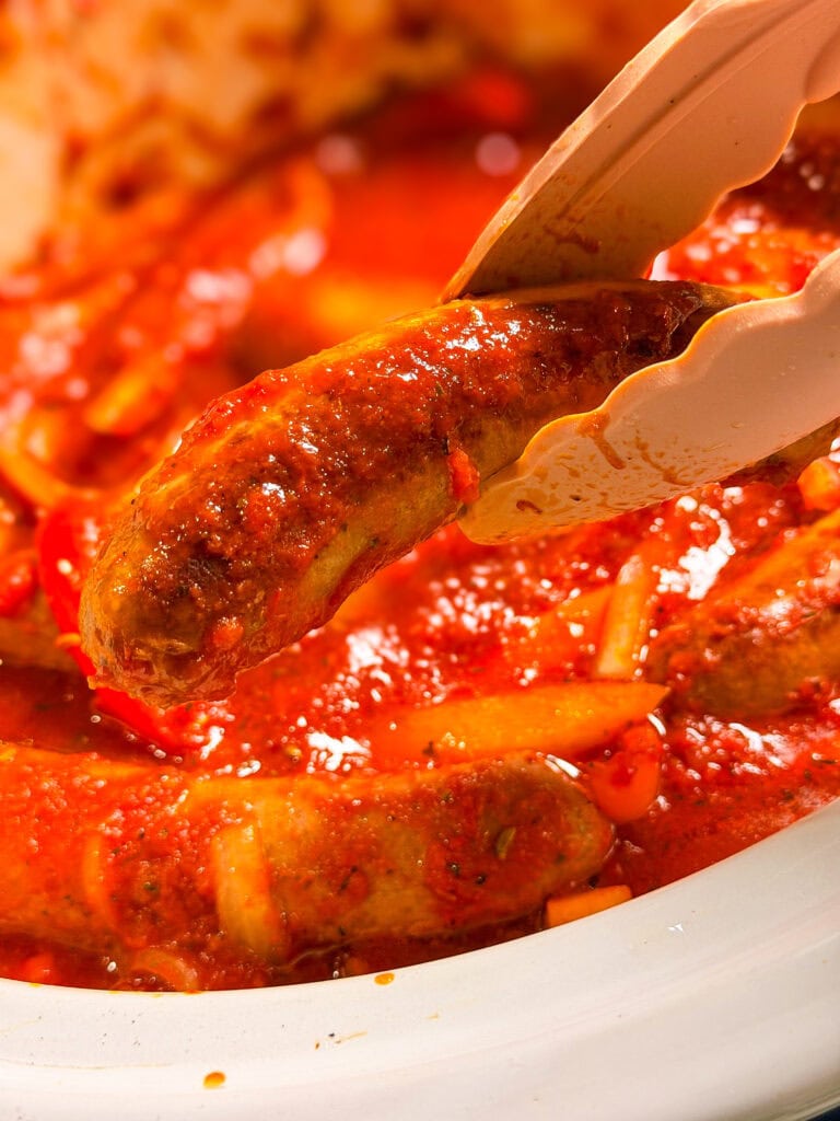 A pair of tongs holds a cooked sausage over a pot of sausages simmering in a tomato-based sauce with sliced peppers and onions.