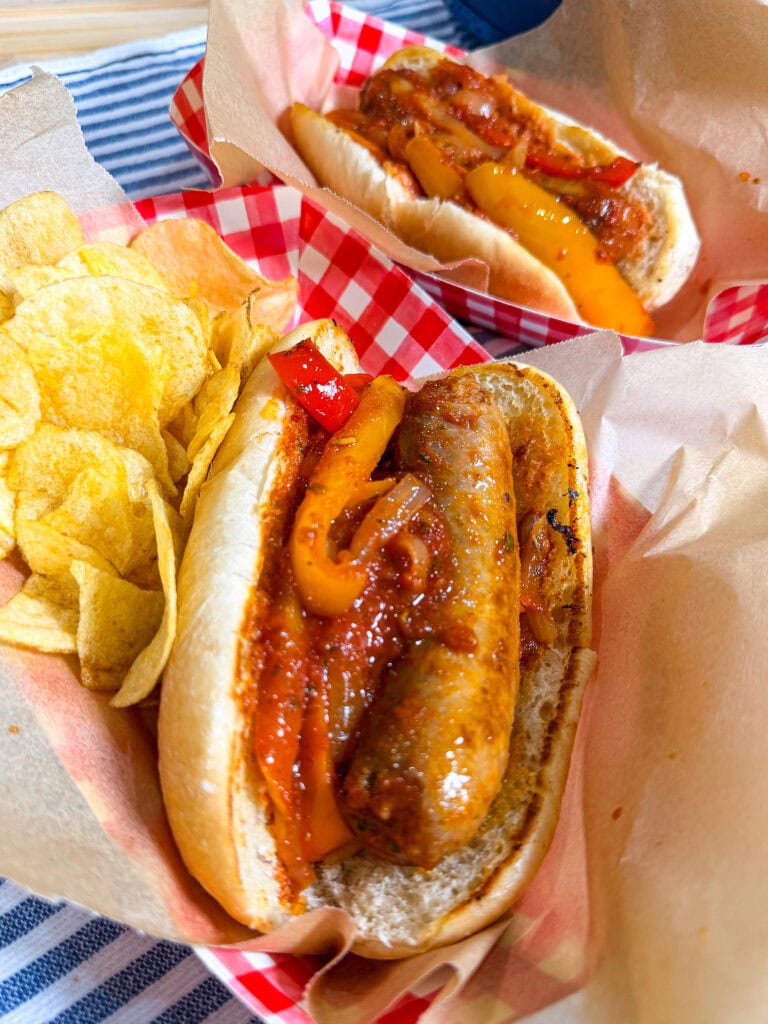 Two sausage sandwiches topped with peppers and sauce are served in paper trays with red checkered liners, next to a side of potato chips.
