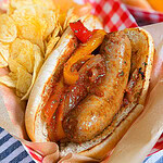 A sausage in a bun with grilled peppers and onions, served with potato chips on the side, all in a red and white paper tray on a blue and white striped cloth.